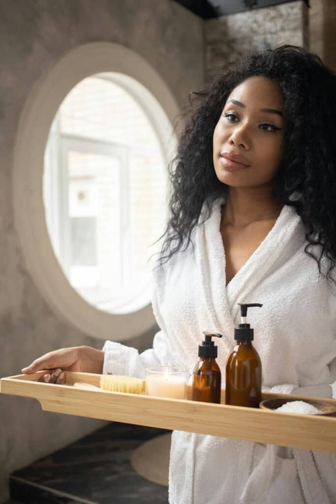 Woman with Afro-textured hair in bathrobe holding wooden tray with shampoo bottles and brush for hair care routine