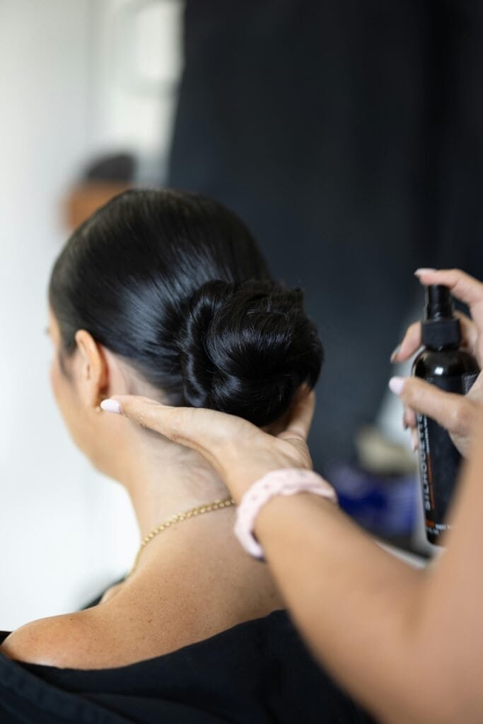 Professional hairstylist holding hair product while styling client's smooth straight black hair pulled into elegant bun