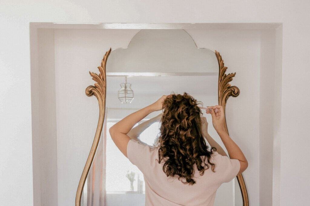 Woman examining her curly hair in ornate mirror while applying natural dandruff treatment to scalp