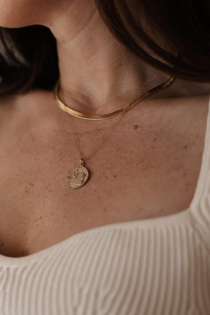 Close-up of a person's neck and chest showing natural hyperpigmentation and sun spots on the skin, particularly visible on the dรฉcolletage area. The person is wearing layered gold necklaces including a snake chain and a coin pendant against a cream-colored ribbed top. This image illustrates how sun exposure commonly affects the chest area, creating the type of hyperpigmentation discussed in our natural treatment guide.