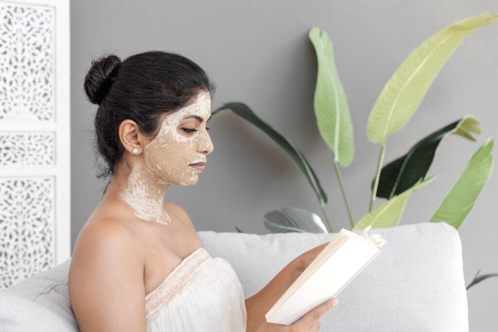 A woman with a natural exfoliating face mask applied to her face and neck, representing gentle removal of pigmented cells as discussed in Pillar 4 of our hyperpigmentation treatment approach. She's relaxing in a white towel while reading, demonstrating how natural treatments can be incorporated into a self-care routine. The mask likely contains ingredients like fruit enzymes, clay, or gentle acids that help with cell turnover and removal of existing hyperpigmentation while being gentle enough to avoid triggering inflammatory responses.