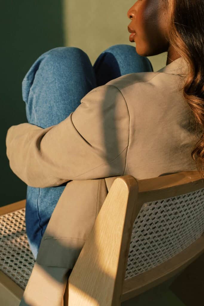 Woman with melanin-rich skin sitting peacefully in a wooden chair, wearing a beige sweater and blue jeans, representing confidence and well-being in skin health management