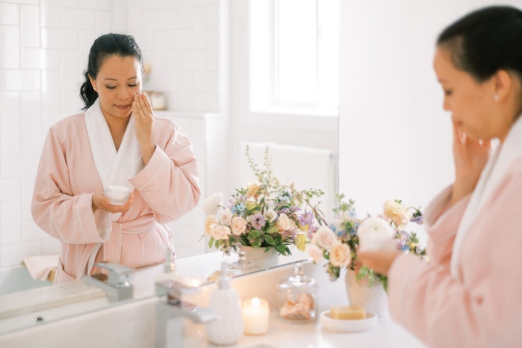 A woman in a pink robe gently applying skincare products to her face while looking in a bathroom mirror, surrounded by candles and fresh flowers. This serene evening scene perfectly captures the dedicated self-care approach of our evening hyperpigmentation repair routine, where active treatment ingredients are applied in a calm, nurturing environment to support overnight skin regeneration and healing.