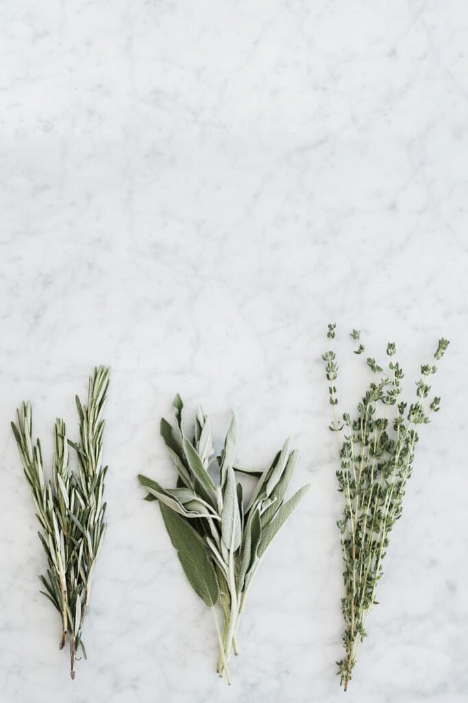 Three bunches of fresh herbs — rosemary, sage, and thyme — on a marble surface, representing polyphenol-rich botanical plants whose antioxidant compounds provide natural photoprotection against UV-induced skin damage