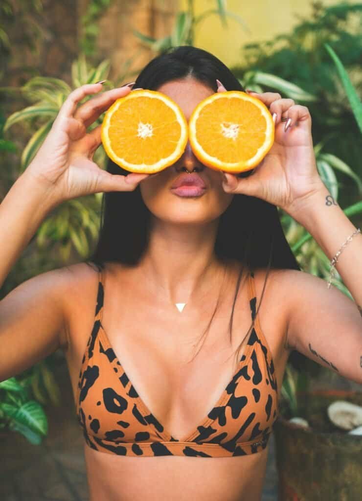 oman holding two halves of a fresh orange over her eyes, playfully illustrating vitamin C's powerful antioxidant role in neutralizing UV-generated free radicals and protecting skin from sun damage and hyperpigmentation