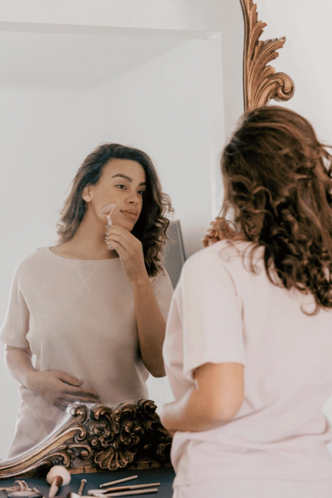 A woman using a jade facial roller tool while looking in an ornate gold mirror, demonstrating self-care and natural skincare practices. This image represents the gentle, consistent approach to treating hyperpigmentation discussed in our step-by-step routine guide, where daily skincare rituals and natural tools support skin health and healing.