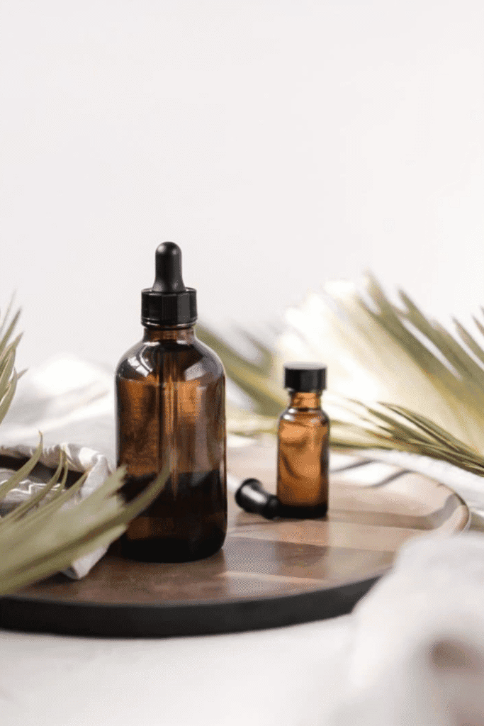 Amber glass dropper bottles containing natural oils arranged on a wooden tray with dried palm leaves, representing the DIY cell renewal oil serum ingredients discussed in our hyperpigmentation routine guide. The dark glass bottles protect light-sensitive oils like rosehip and sea buckthorn extracts, essential components for creating your own barrier repair and pigmentation-fading serum at home.