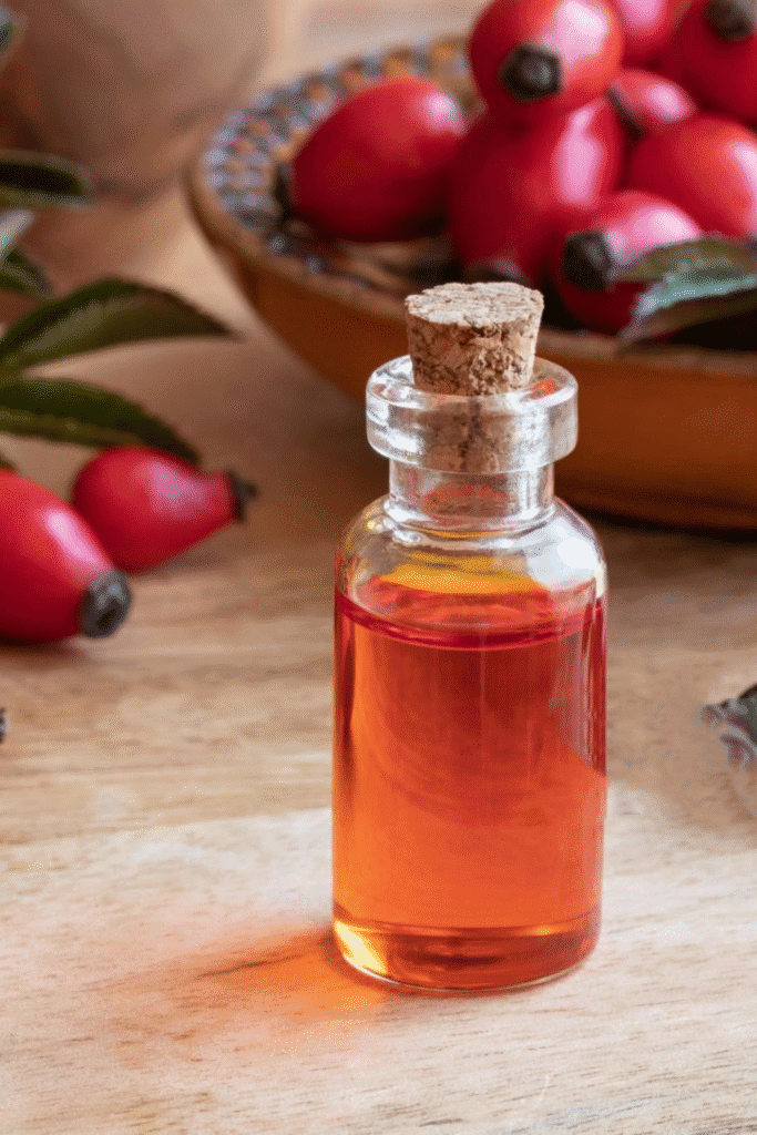 Clear glass bottle of golden rosehip oil with cork stopper beside a bowl of fresh rosehips, illustrating this natural sunburn treatment oil rich in vitamins A and E for skin repair and restoration