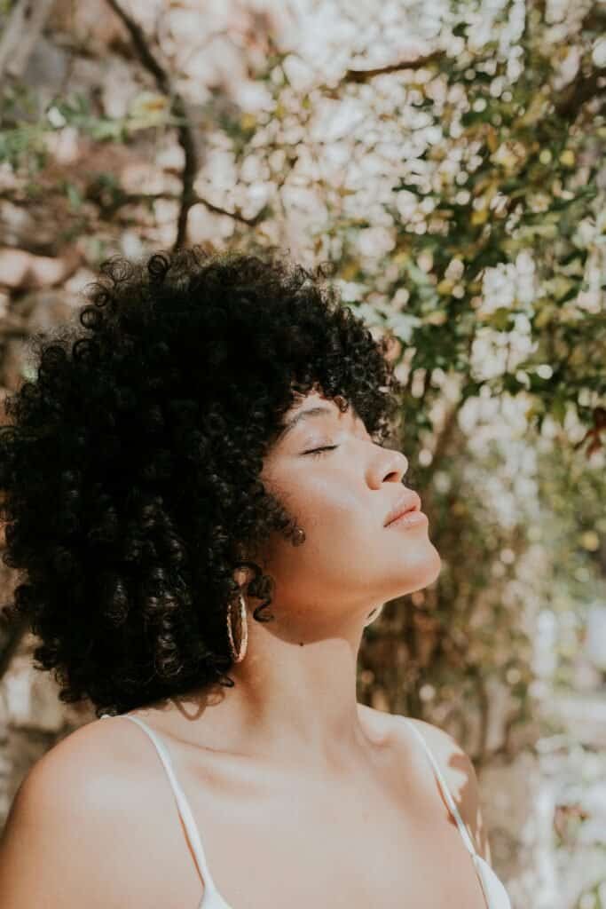 Black woman with natural curly afro hair and eyes closed, face lifted toward dappled sunlight through trees, representing low photosensitivity skin type VI with high melanin content and strong natural UV protection