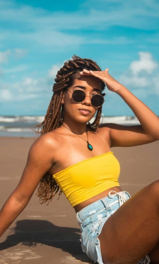 Melanin-rich Black woman with braids and sunglasses shielding her eyes from the sun at the beach, illustrating how different skin phototypes respond differently to UV radiation and photosensitivity
