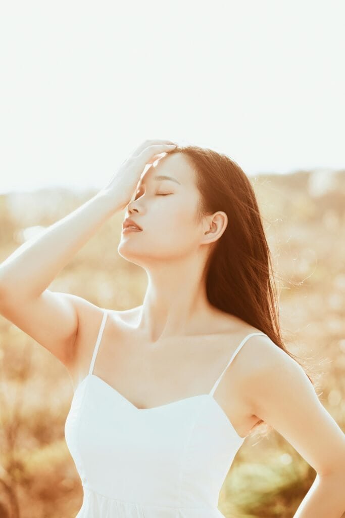 Asian woman with fair skin shielding her face from intense sunlight with closed eyes, representing moderate photosensitivity and the skin's natural protective response to UV radiation exposure