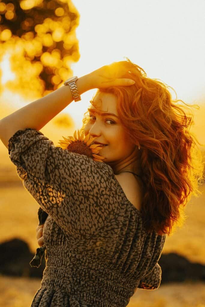 Fair-skinned red-haired woman holding a sunflower in warm golden sunset light, representing Fitzpatrick skin type I-II with high photosensitivity and greater vulnerability to UV-induced sun damage