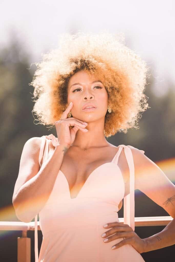 Light-skinned Black woman with voluminous blonde afro standing in bright sunlight, illustrating the spectrum of melanin levels and varying sun protection needs across different Black skin tones