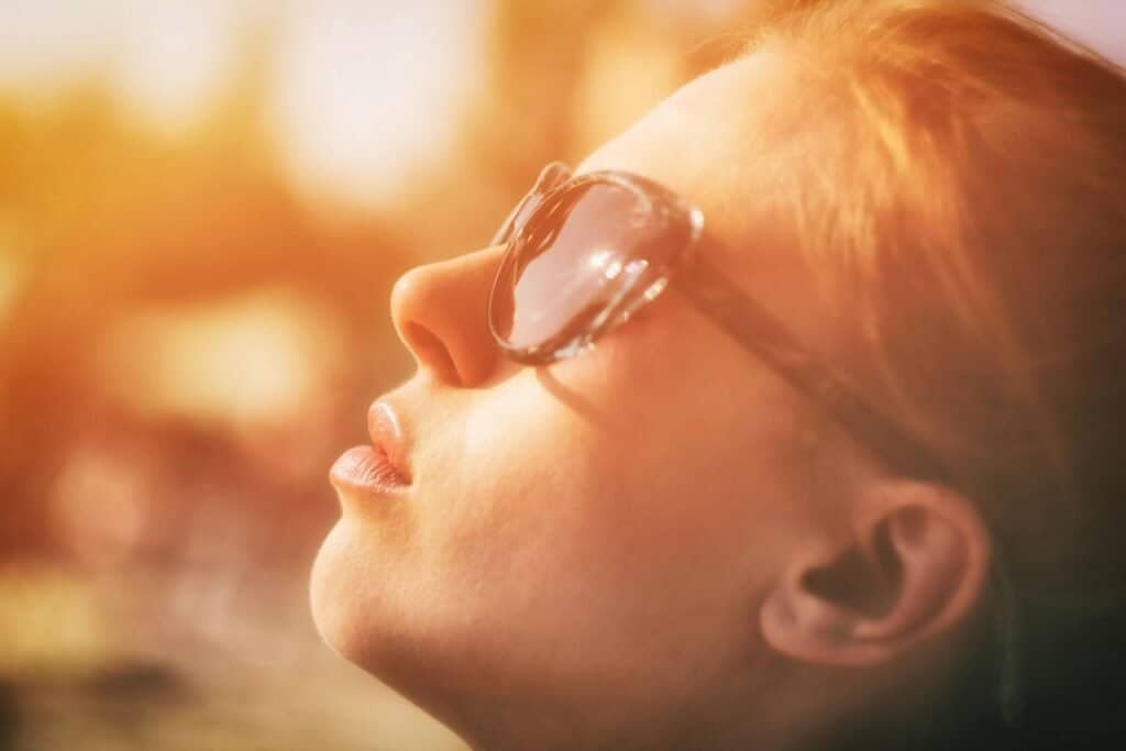 Close-up of a fair-skinned red-haired young woman wearing sunglasses looking up toward intense sunlight, illustrating how sunscreen use in high-risk Fitzpatrick skin types can suppress essential vitamin D synthesis