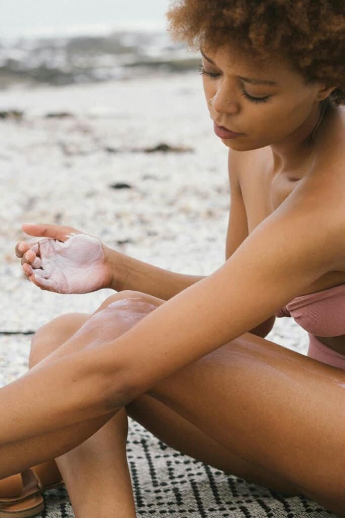Black woman in bikini applying sunscreen to her legs at the beach, highlighting the gap in sunscreen testing that excludes darker skin tones despite universal marketing of SPF products