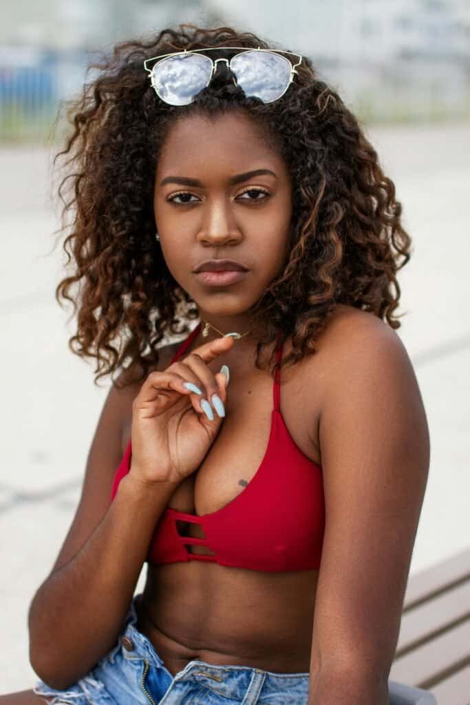 Confident Black woman with curly hair and sunglasses at the beach, representing melanin-rich skin types excluded from SPF testing despite being targeted by universal sunscreen marketing campaigns