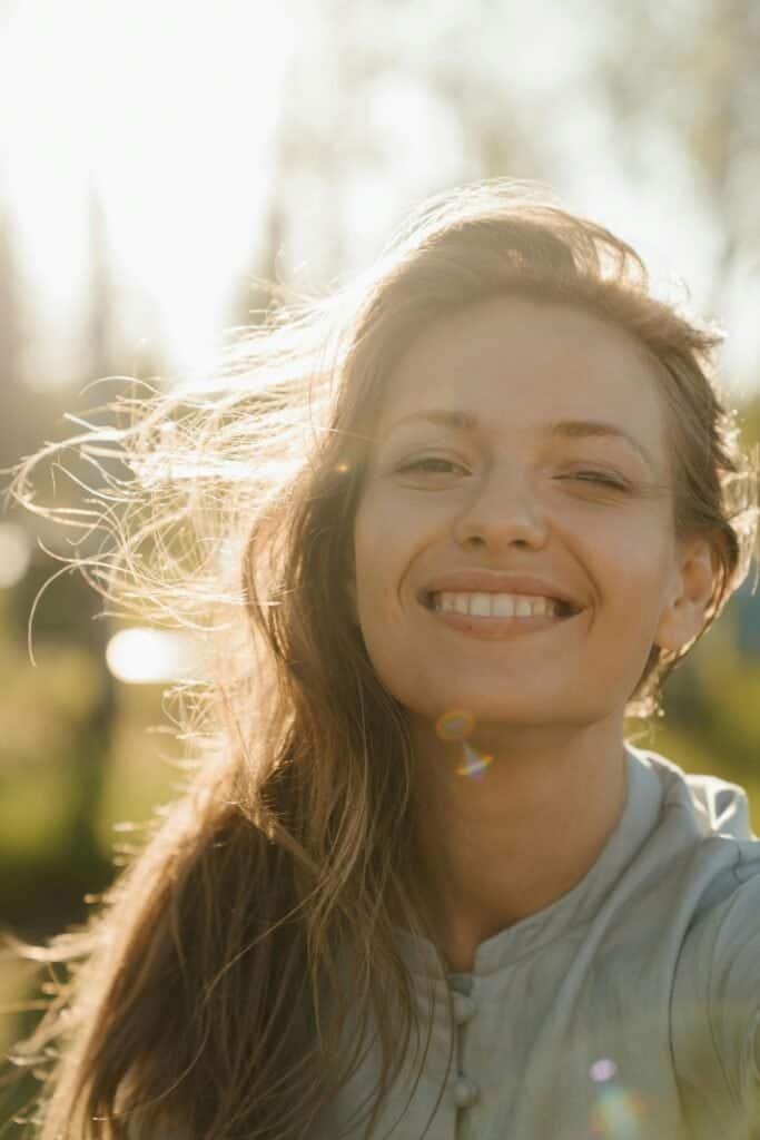 Smiling fair-skinned woman with windswept hair bathed in warm sunlight, representing the balanced approach to sun safety beyond sunscreen including antioxidants and physical UV protection