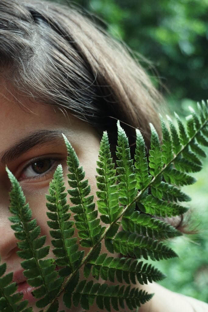 Close-up of woman's face behind fresh fern fronds representing Polypodium leucotomos for skin health and anti-aging