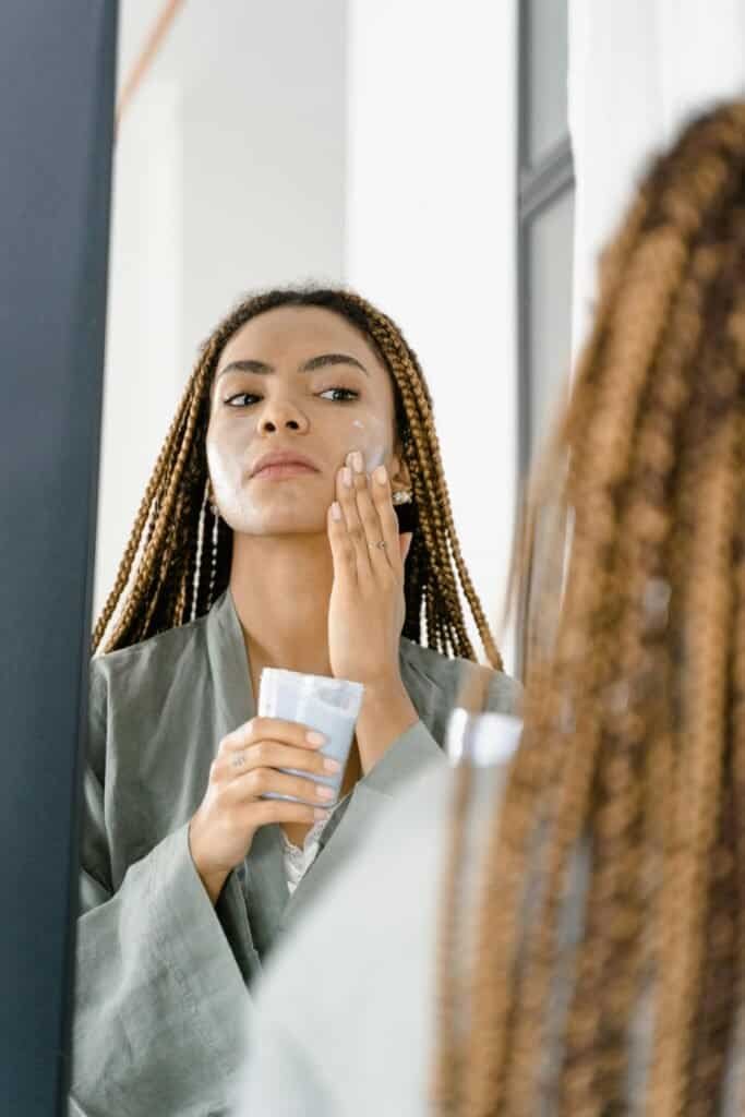 Black woman with braids applying skincare cream to her face in a mirror, representing an acne-aware skincare approach for women with acne-prone skin