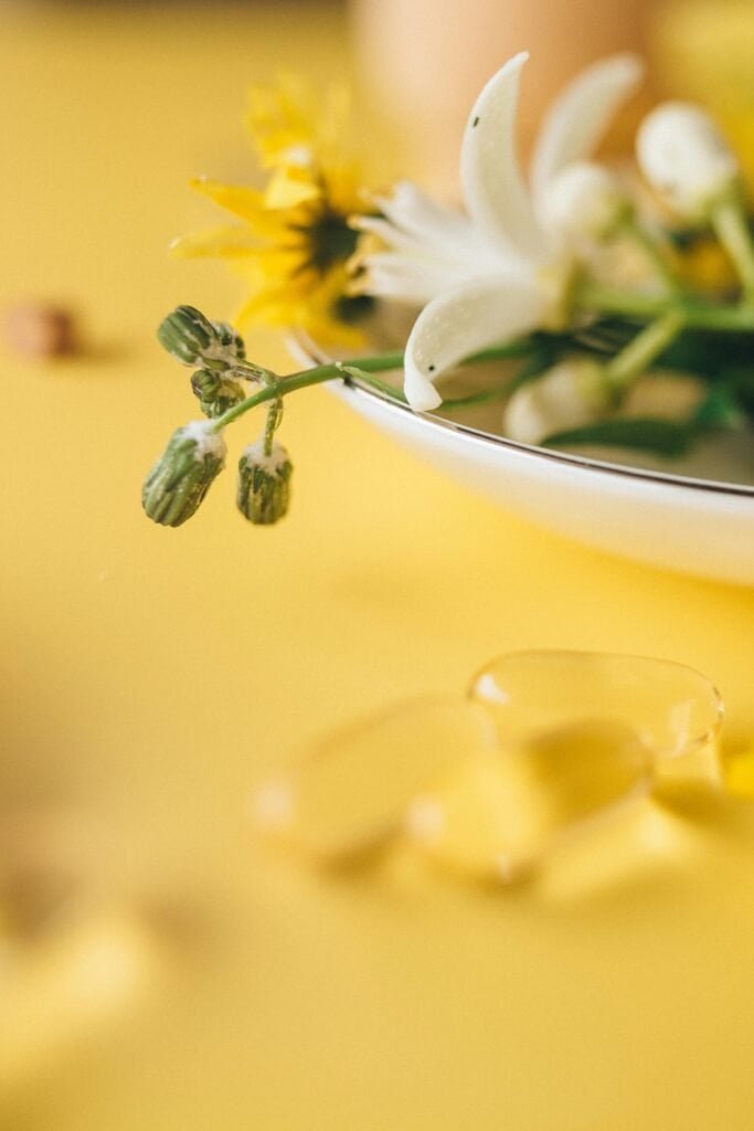 Botanical plant buds and white flowers in a white bowl alongside gel supplement capsules on a yellow background, illustrating the topical and oral routes of botanical ingredients for acne
