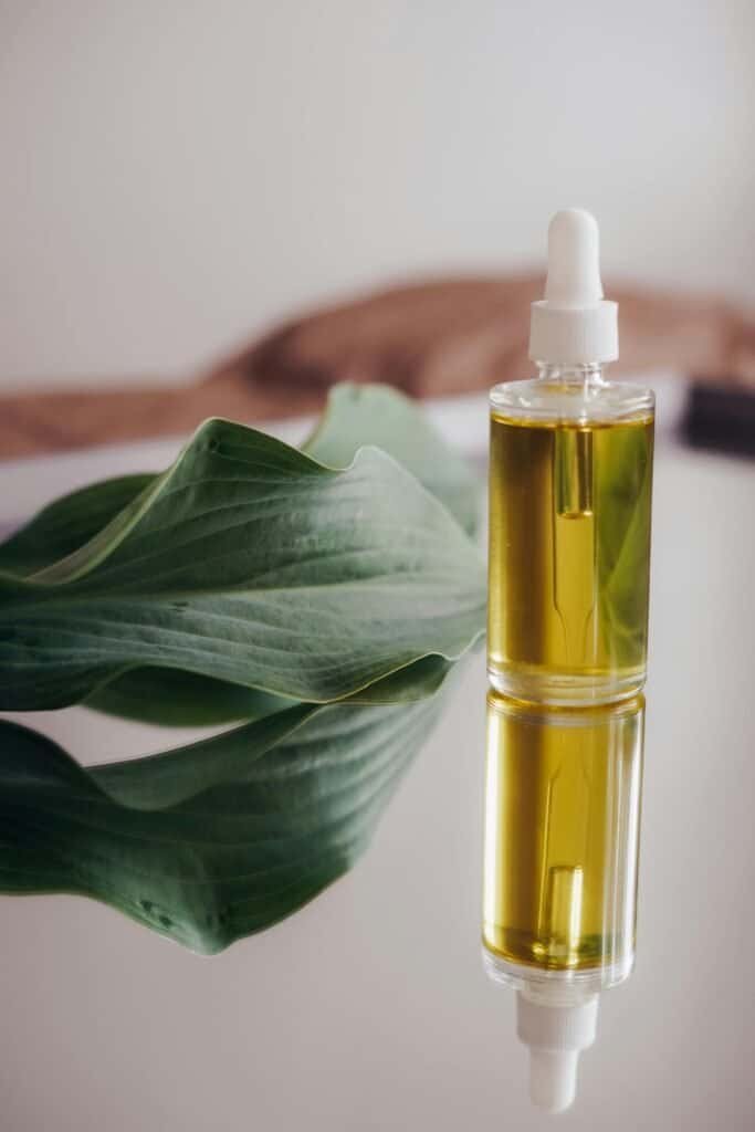 Clear glass dropper bottle filled with golden botanical facial oil beside a large green tropical leaf on a reflective white surface, representing the DIY retinoid-rich rosehip and sea buckthorn facial oil for acne-prone skin