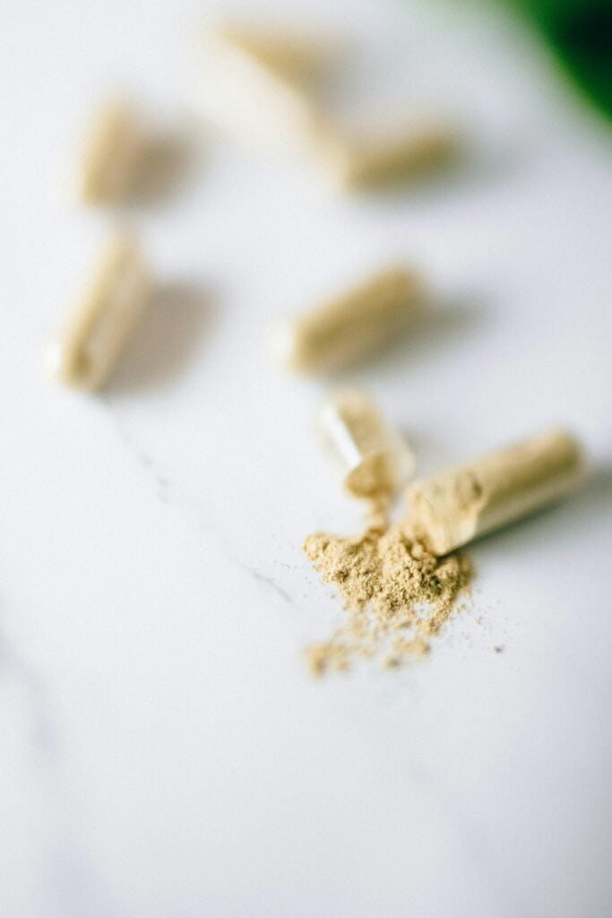 Open supplement capsule spilling fine botanical powder onto a white marble surface surrounded by other capsules, representing targeted nutraceutical supplementation for acne including zinc picolinate and selenium