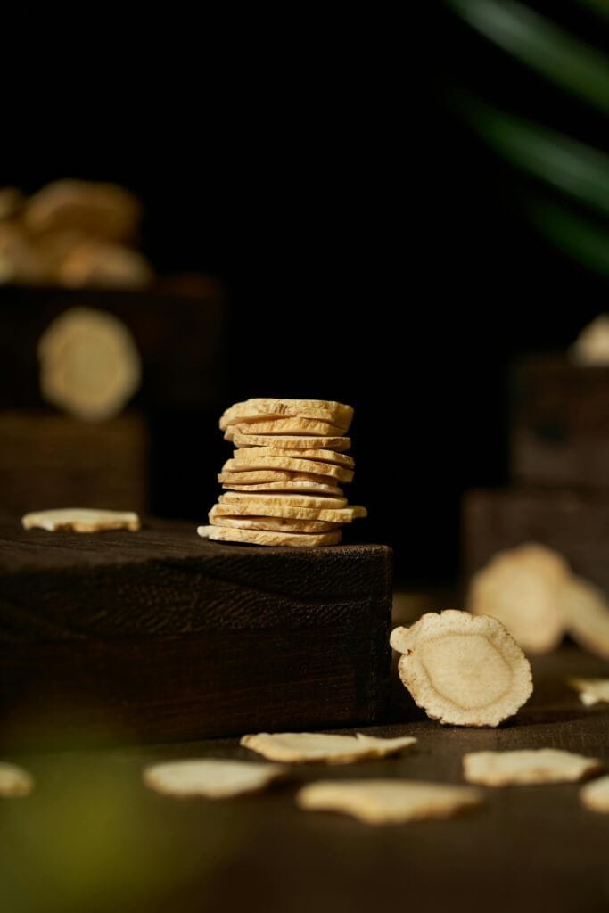 Stack of dried licorice root slices on a dark wooden block with scattered pieces, representing the Glycyrrhiza root from which glycyrrhetinic acid and licochalcone A are extracted for acne and hyperpigmentation treatment