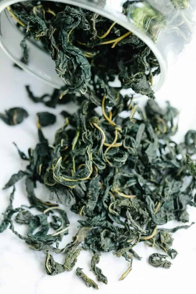 Dried green tea leaves spilling from a glass container onto a white surface, representing EGCG green tea extract and its clinically studied anti-acne and sebum-reducing properties
