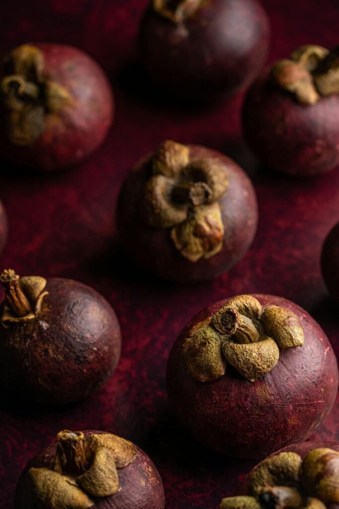 Close-up of whole mangosteen fruits with their distinctive calyxes on a deep red background, representing the queen of fruits whose pericarp contains potent anti-acne xanthone compounds