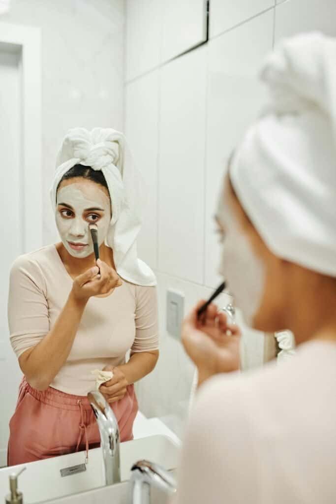 Woman with a towel on her head applying a white clay face mask with a brush in front of a bathroom mirror, demonstrating the application technique of a DIY clay mask for acne-prone skin