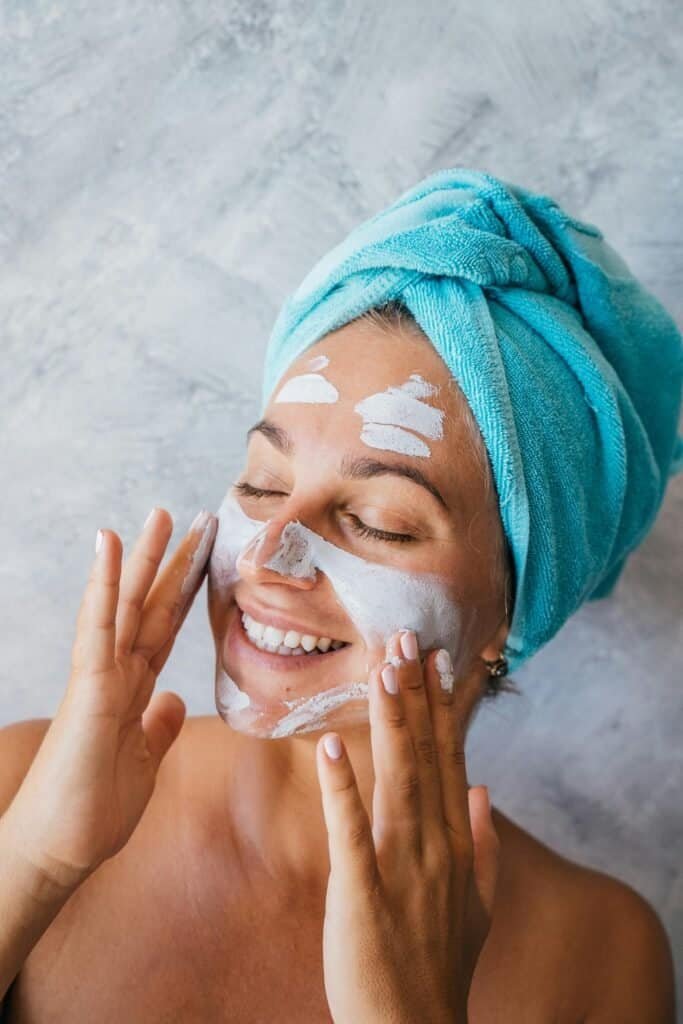 Smiling woman with a blue towel on her head applying a white clay cleanser to her face with her hands, illustrating the cleansing step of an acne-prone skin routine