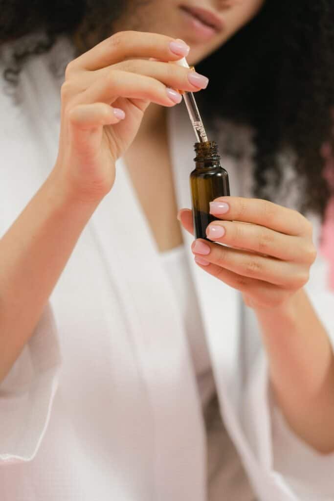 Woman with curly hair in a white robe dispensing drops from a small amber glass dropper bottle, showing how to apply the DIY retinoid-rich facial oil for acne-prone skin moisturizing and repair