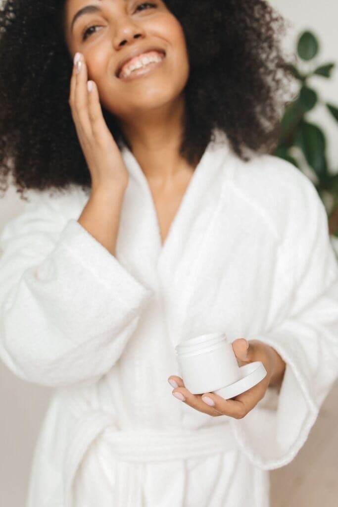 Smiling woman applying moisturizer to her face while holding an open jar of cream