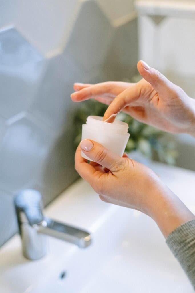 Close-up of hands scooping moisturizer from a jar at a bathroom sink